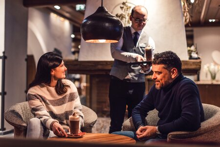 Zwei Gäste sitzen bei Getränken unter einer Hängelampe in einem gemütlichen Restaurantbereich, während ein Kellner im Hintergrund serviert.