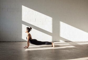 A woman holds a cobra yoga pose in a bright room with large patches of light on the wall.