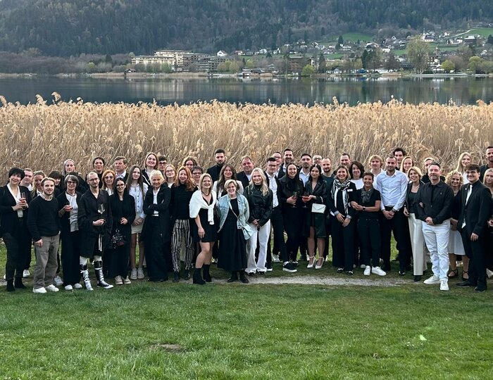 Eine große Gruppe von Gästen steht gemeinsam auf einer Wiese vor dem See mit Schilf und Bergen im Hintergrund.