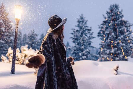 Eine Frau steht im tief verschneiten Winterwald neben einer Laterne und blickt in die Schneelandschaft.