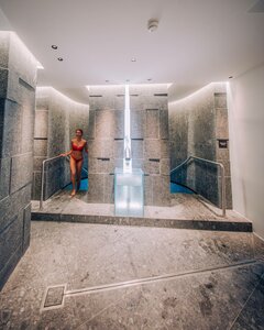 Bilder vom Wellnessurlaub mit Abwechslung in Österreich. Eine Frau in einem roten Badeanzug steht in einem modernen Spa-Bereich mit grauen Steinwänden. Der Raum ist gut beleuchtet und hat eine entspannende Atmosphäre. Im Hintergrund sind Wasserbecken und Handläufe sichtbar.