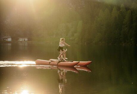 Eine Person paddelt auf einem SUP-Board über den ruhigen See vor bewaldeten Hängen im Abendlicht.