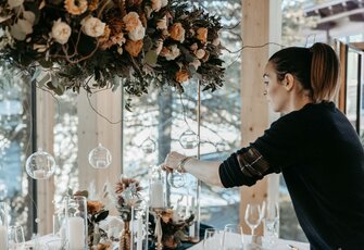 A woman decorates a festively set table with floral arrangements and glass ornaments.