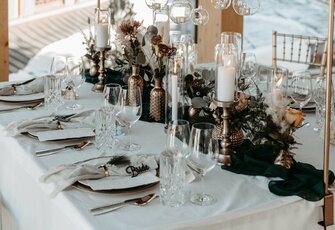 A festive table setting with a white tablecloth, glasses, candles, and lush floral decoration by the window.