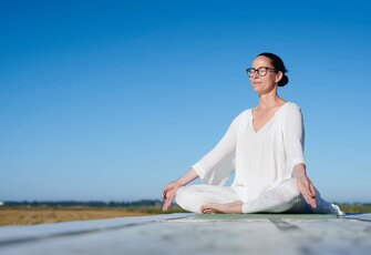 A woman sits cross-legged on a platform by the lakeshore under a clear blue sky.