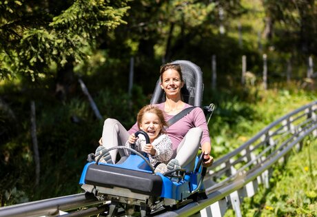 Eine Frau und ein Kind fahren gemeinsam auf einer Sommerrodelbahn durch die grüne Berglandschaft.