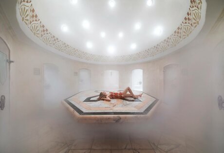 A person lies relaxed on a raised stone platform in the hamam, surrounded by warm steam and soft light.