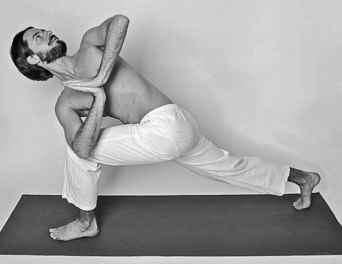 Ein Mann zeigt eine tiefe Yoga-Position auf einer Matte vor einer schlichten Wand.