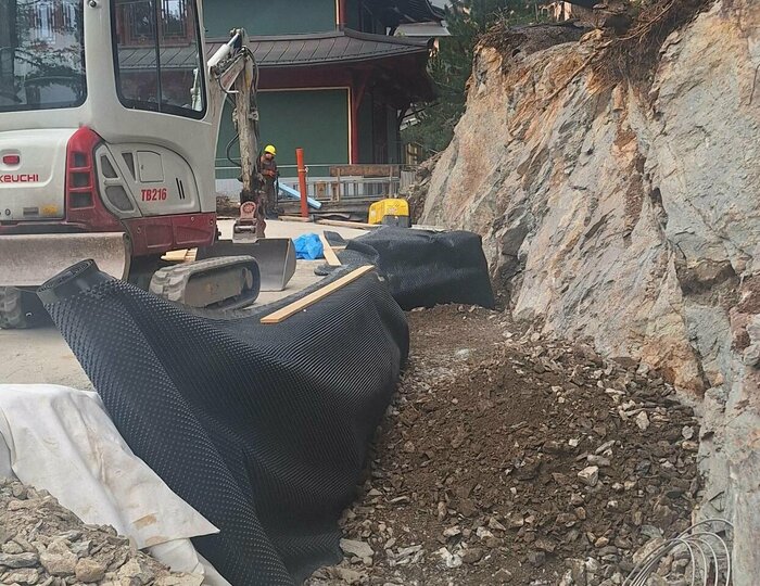 Baustelle am Hotel Hochschober in Woche 5 Ein Minibagger arbeitet neben dem Hotelgebäude an einer Baugrube mit Steinmaterial und Abdeckplanen.