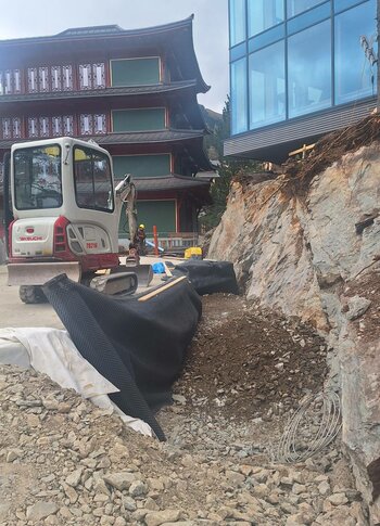Baustelle am Hotel Hochschober in Woche 5 Ein Minibagger arbeitet neben dem Hotelgebäude an einer Baugrube mit Steinmaterial und Abdeckplanen.