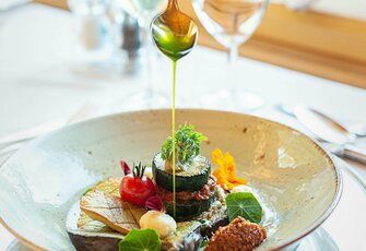 Eine Hand beträufelt ein kunstvoll angerichtetes Gourmetgericht mit Sauce auf einem gedeckten Tisch.