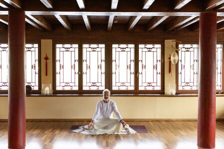 Eine Frau sitzt in weißer Kleidung auf einer Yogamatte in einem ruhigen Raum mit Holzdecke und Fenstern im Chinaturm.
