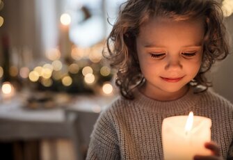 Ein Kind hält eine brennende Kerze und blickt lächelnd nach unten, im Hintergrund leuchten weiche Weihnachtslichter.