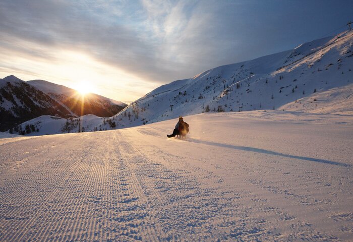 Eine Person fährt auf einer verschneiten Piste im Sonnenaufgang, umgeben von winterlichen Bergen.