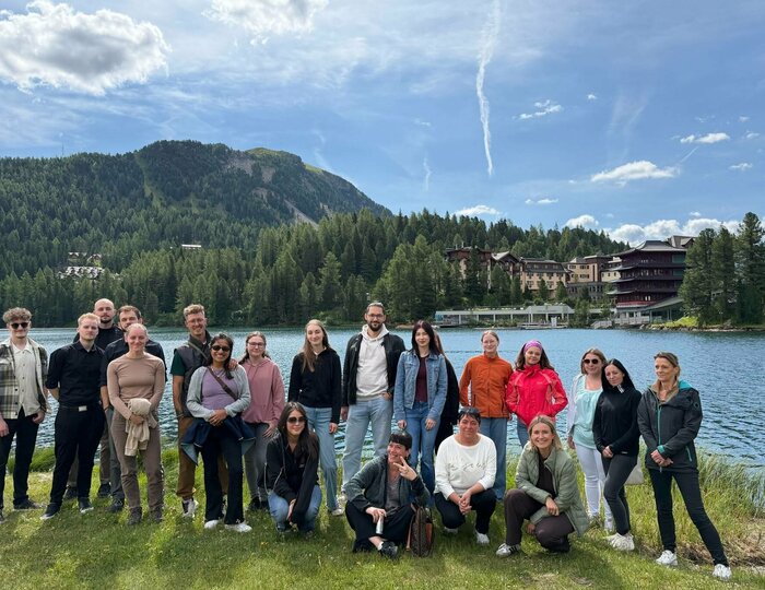 Eine Gruppe von Menschen steht und sitzt am Ufer des Sees vor dem Hotel Hochschober und bewaldeten Bergen.