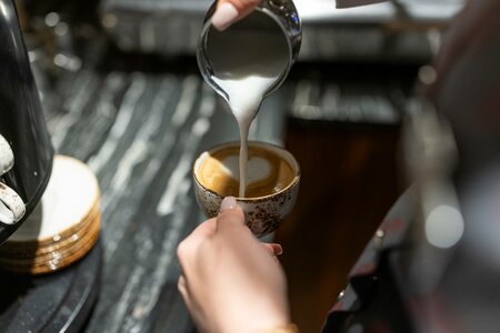 Eine Hand gießt Milch aus einer Kanne in eine kleine Tasse Kaffee.