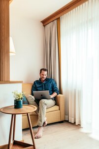 Ein Mann sitzt in einem Sessel im hellen Hotelzimmer und arbeitet am Laptop neben dem Fenster.
