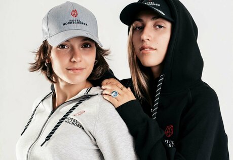 Two young women wear hoodies and caps with the Hotel Hochschober logo against a light background.