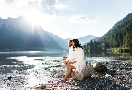 Ein Paar sitzt auf einem Felsen am Ufer des Sees und blickt in die sonnige Berglandschaft.