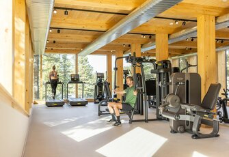 Ein Mann trainiert an einem Kraftgerät im hellen Fitnessraum mit Holzdecke und Blick ins Grüne.