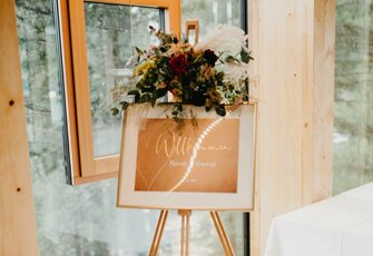A floral arrangement decorates a welcome sign on an easel in a bright room.