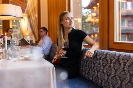 Eine Frau sitzt im Hotelrestaurant am Fenster und hält ein Glas Wein, während im Hintergrund weitere Gäste speisen.