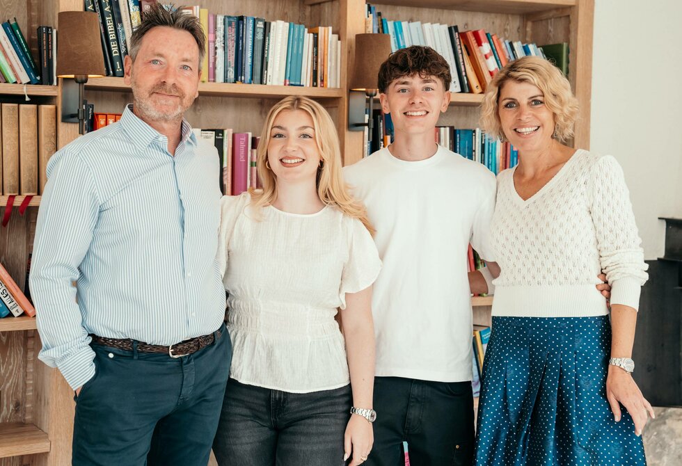 The Leeb and Klein family stands in front of a bookshelf at Hotel Hochschober and looks at the camera.