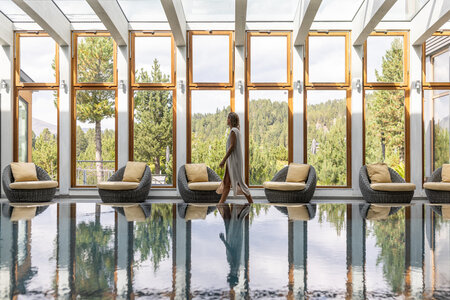 Eine Frau geht an gepolsterten Loungesesseln am Pool vorbei, vor großen Fenstern mit Blick ins Grüne.