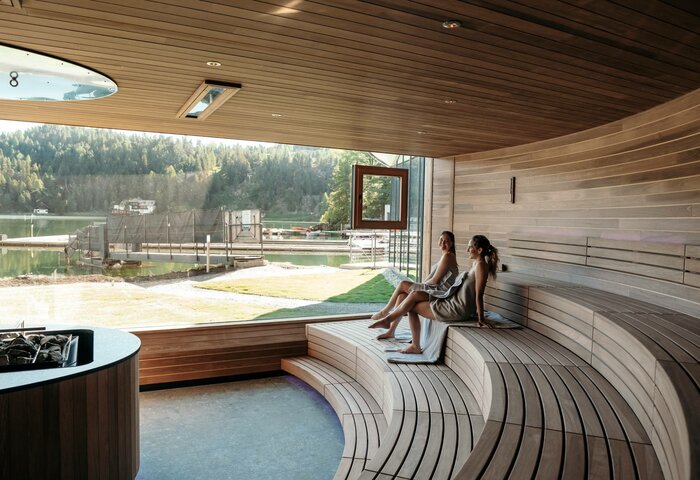 Two women sit in the panorama sauna overlooking the lake and the wooded mountain landscape.