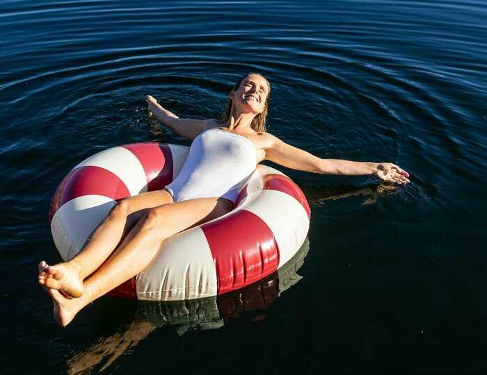 Urlaubsangebote für das ganze Jahr - Turracher Höhe Eine Frau in einem weißen Badeanzug entspannt auf einem rot-weißen Schwimmring in einem ruhigen See. Umgeben von grünen Bäumen und sanften Hügeln spiegelt sich die Landschaft im Wasser. Die Atmosphäre ist friedlich und sonnig.