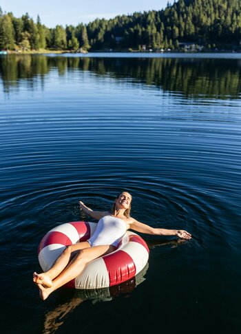 Eine Frau in einem weißen Badeanzug entspannt auf einem rot-weißen Schwimmring in einem ruhigen See. Umgeben von grünen Bäumen und sanften Hügeln spiegelt sich die Landschaft im Wasser. Die Atmosphäre ist friedlich und sonnig.