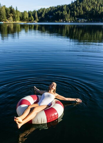 Urlaubsangebote für das ganze Jahr - Turracher Höhe Eine Frau in einem weißen Badeanzug entspannt auf einem rot-weißen Schwimmring in einem ruhigen See. Umgeben von grünen Bäumen und sanften Hügeln spiegelt sich die Landschaft im Wasser. Die Atmosphäre ist friedlich und sonnig.