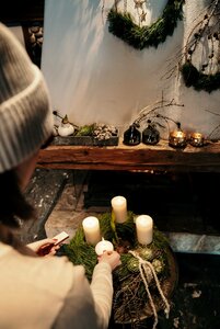Eine Person dekoriert einen Adventskranz mit Kerzen und Tannengrün vor einer rustikalen Wand.