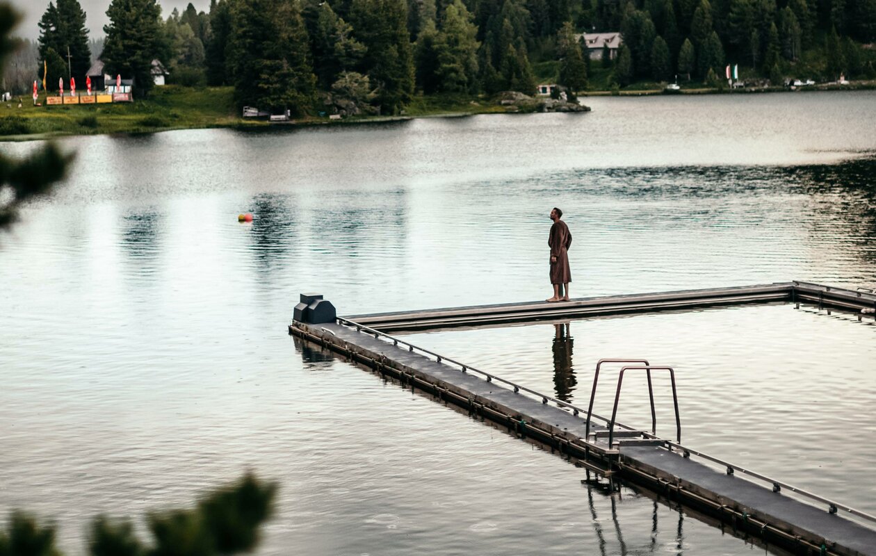 Eine Person steht auf dem Steg des beheizten See-Bads im Turracher See, umgeben von ruhigem Wasser und Uferwald.