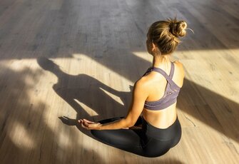 A woman sits in a yoga pose on the wooden floor, seen from behind in warm light.