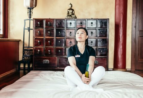 Eine Mitarbeiterin sitzt auf einem Bett vor einem Schrank mit vielen kleinen Fächern und hält eine Flasche in den Händen.