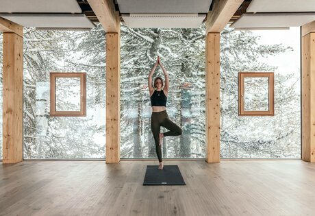 A woman practices yoga on a mat in a bright room with a view of the snowy forest.