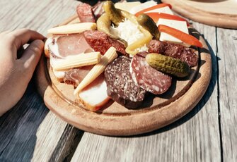 Eine Hand greift neben einer rustikalen Vorspeisenplatte mit Käse, Wurst, Gurken und Brot auf einem Holztisch.