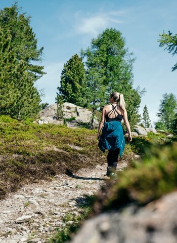 Eine Frau wandert auf einem sonnigen Bergweg zwischen Wiesen und Felsen.