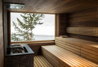 Ein heller Saunaraum mit Holzverkleidung und einer großen Fensterfront, die einen Blick auf schneebedeckte Landschaften und einen Baum bietet. Die Bänke sind aus hellem Holz, und ein Steingrill steht in der Ecke.