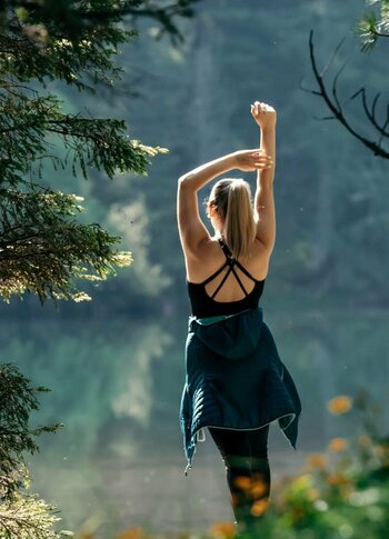 Eine Frau übt Yoga am Ufer des Sees, umgeben von Bäumen und sommerlicher Natur.