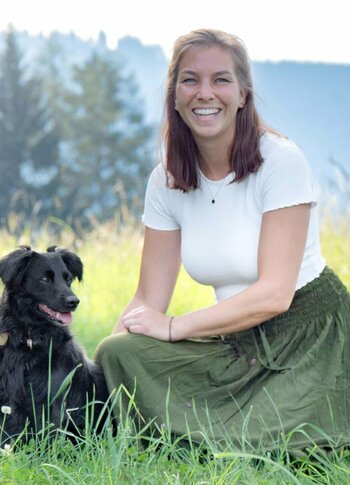 Eine Frau sitzt mit einem schwarzen Hund im hohen Gras vor einer Bergkulisse.