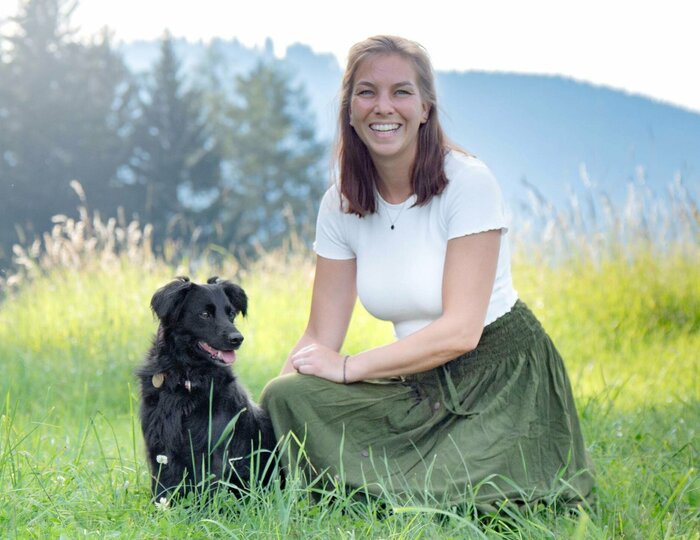 Hundetraining mit Frau und Hund im Grünen Eine Frau sitzt mit einem schwarzen Hund im hohen Gras vor einer Bergkulisse.