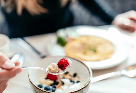 Eine Hand hält einen Löffel mit Himbeeren über einer Müslischale beim Frühstück im Hotelrestaurant.