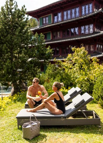 Ein Paar sitzt entspannt auf Liegen im Freien, umgeben von grünen Pflanzen und Bäumen. Sie genießen Cocktails und unterhalten sich. Im Hintergrund ist ein großes, traditionelles Gebäude mit mehreren Etagen sichtbar.
