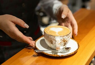 Eine Person stellt eine Tasse Cappuccino mit Herzmuster auf einem Tisch ab.