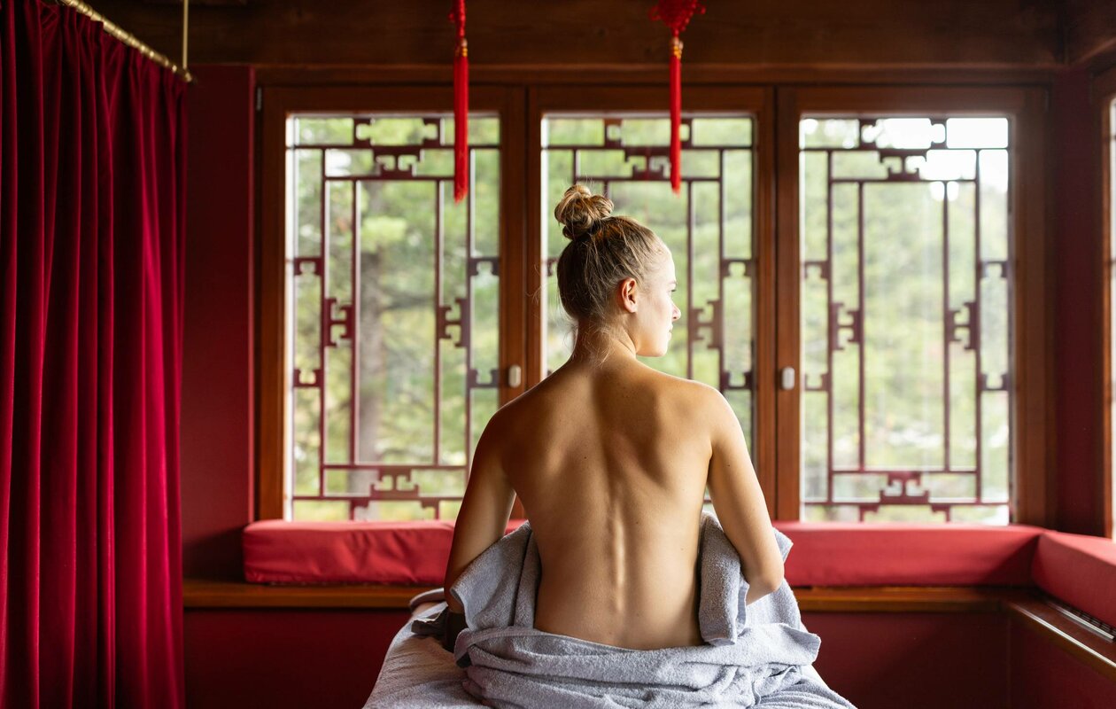 Eine Frau sitzt mit freiem Rücken auf einer Liege im Chinaturm vor chinesisch inspirierten Fenstern.