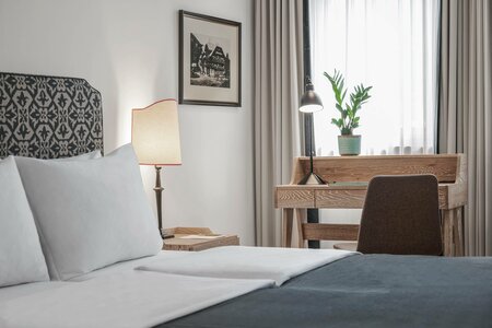 Ein helles Hotelzimmer mit Bett, Schreibtisch, Stuhl, Stehlampe und Fenster mit Vorhängen.