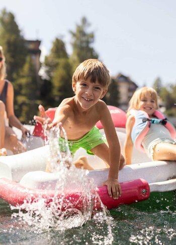 A woman sits on a floating platform in a lake, smiling at two children playing. A boy in green swim trunks splashes water as he leans over the edge, while a girl in a life jacket relaxes beside him. Lush trees and buildings are visible in the background.