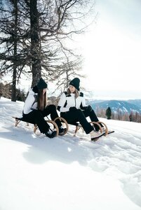 Zwei Frauen sitzen auf Holzschlitten im Schnee mit Blick auf die Winterlandschaft auf der Turracher Höhe.